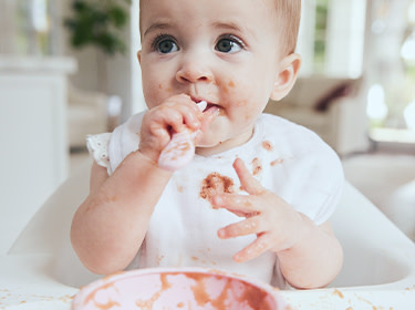 A child with food stains on his face and clothes sits next to the table, holding a spoon in his hands