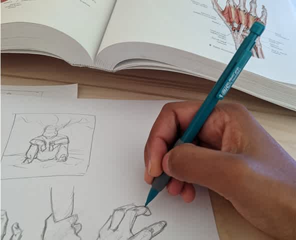 Artist Hillary Wilson sketches on a piece of paper with a mechanical pencil. A medical book is open for reference displaying an anatomical image of a hand.