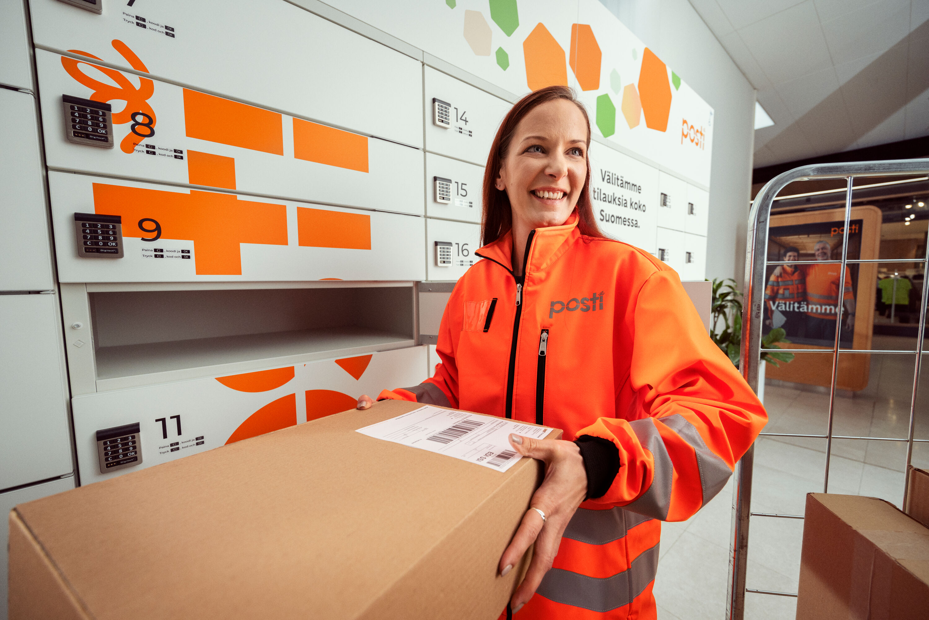 Hymyilevä työntekijä Postin oranssissa takissa kantaa pakettia.