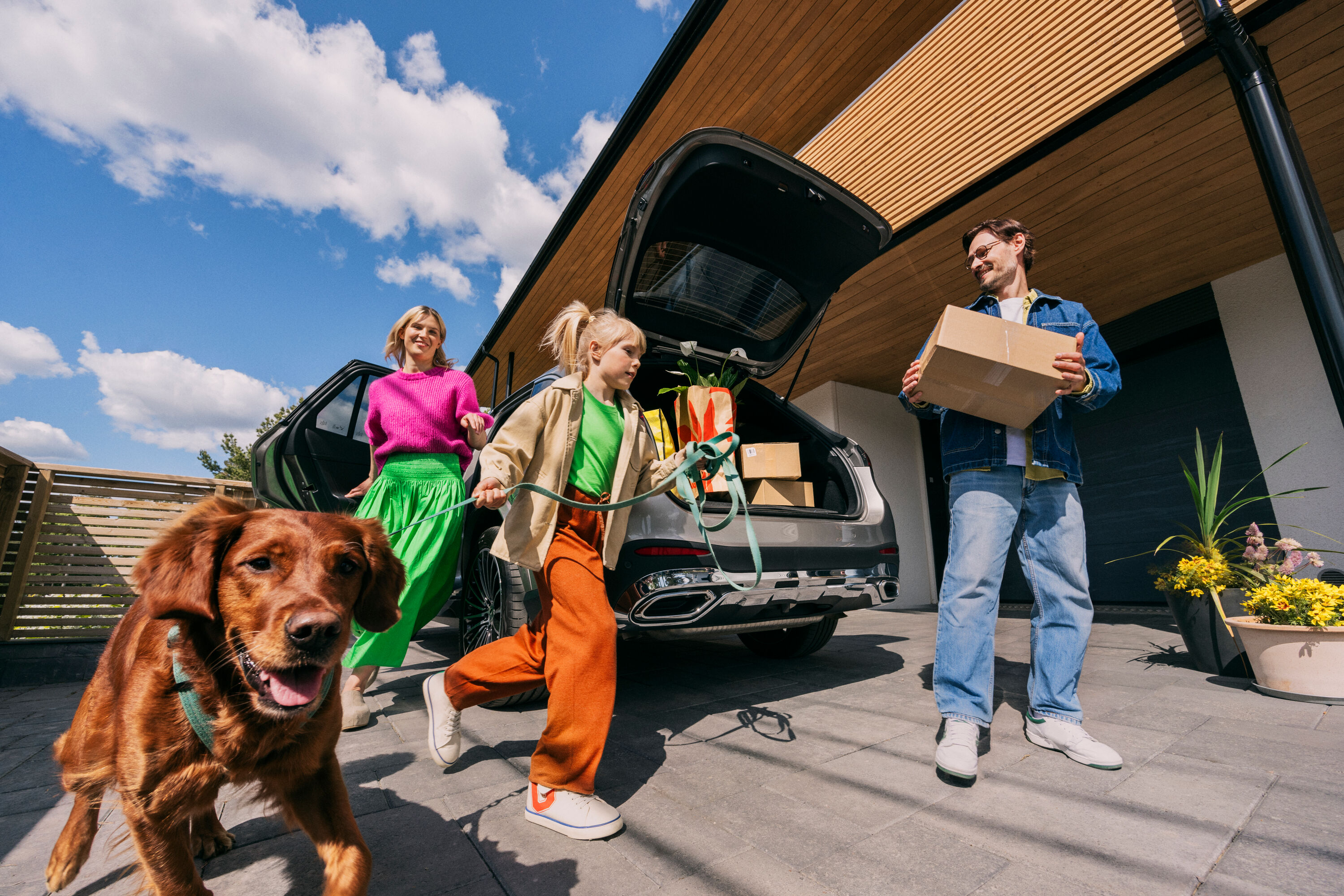 En familj lastar ur saker från bilens bagageutrymme framför huset. En hund är med.