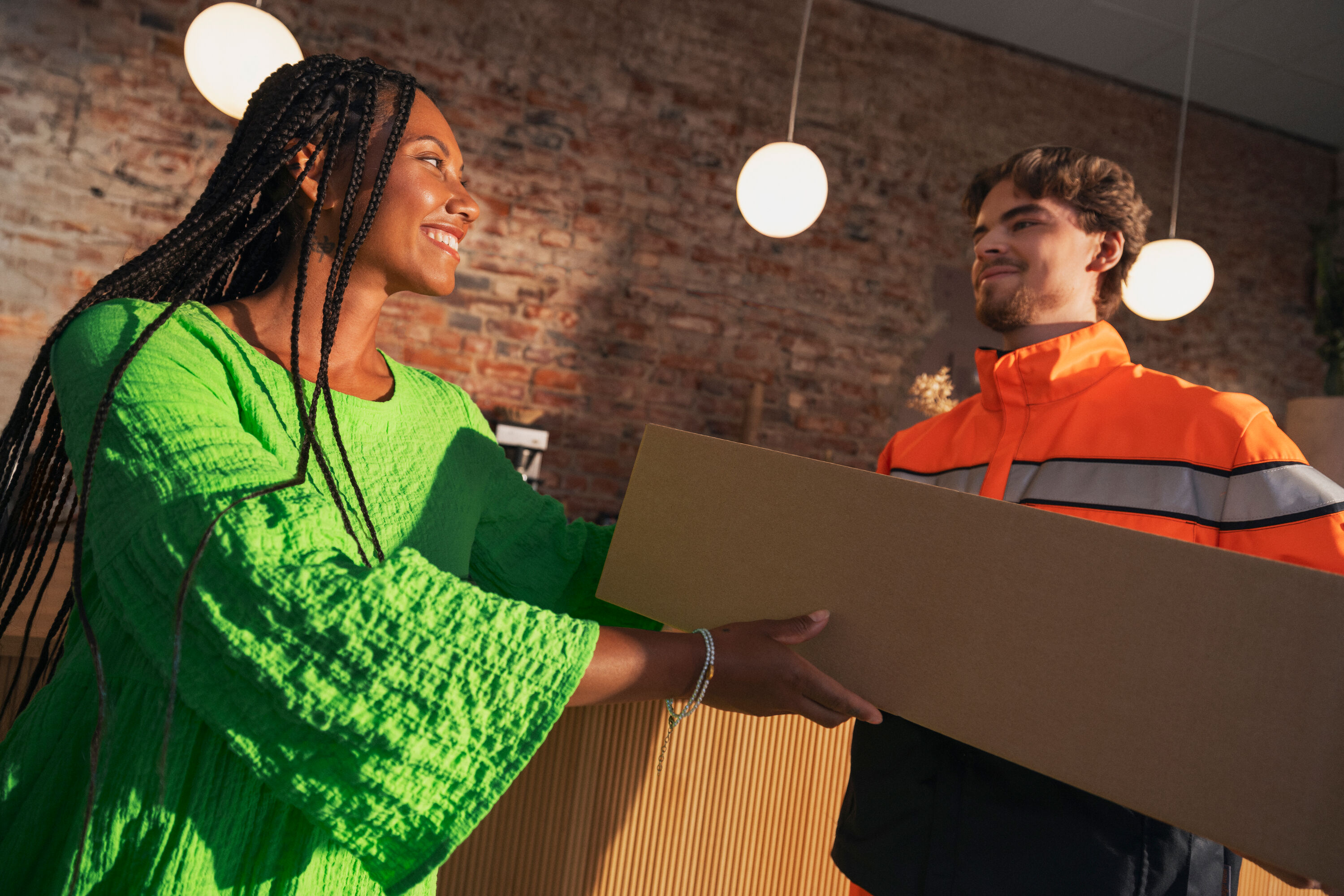 En kvinna i grön tröja tar emot ett paket från en Posti-anställd med orange jacka.
