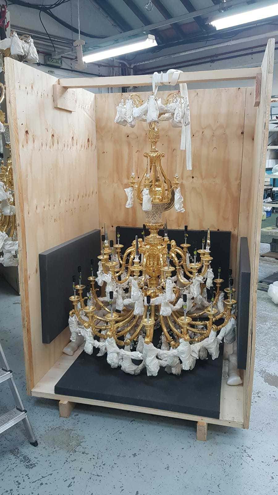 A large, ornate gold chandelier is shown secured inside a custom wooden crate. Various parts are wrapped in protective white padding, and it's mounted against a black backboard. The photo appears to be taken in a workshop or storage area.