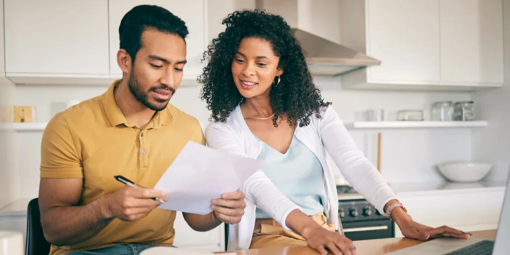Couple reviewing documents together at kitchen table with laptop, man in yellow shirt holding paper, woman in white top smiling.