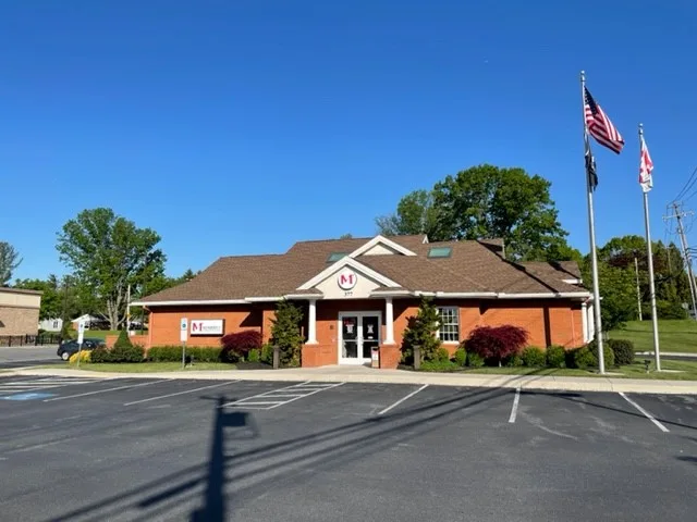 Orange brick building with brown roof, white columns, American flag, and parking lot under clear blue sky.