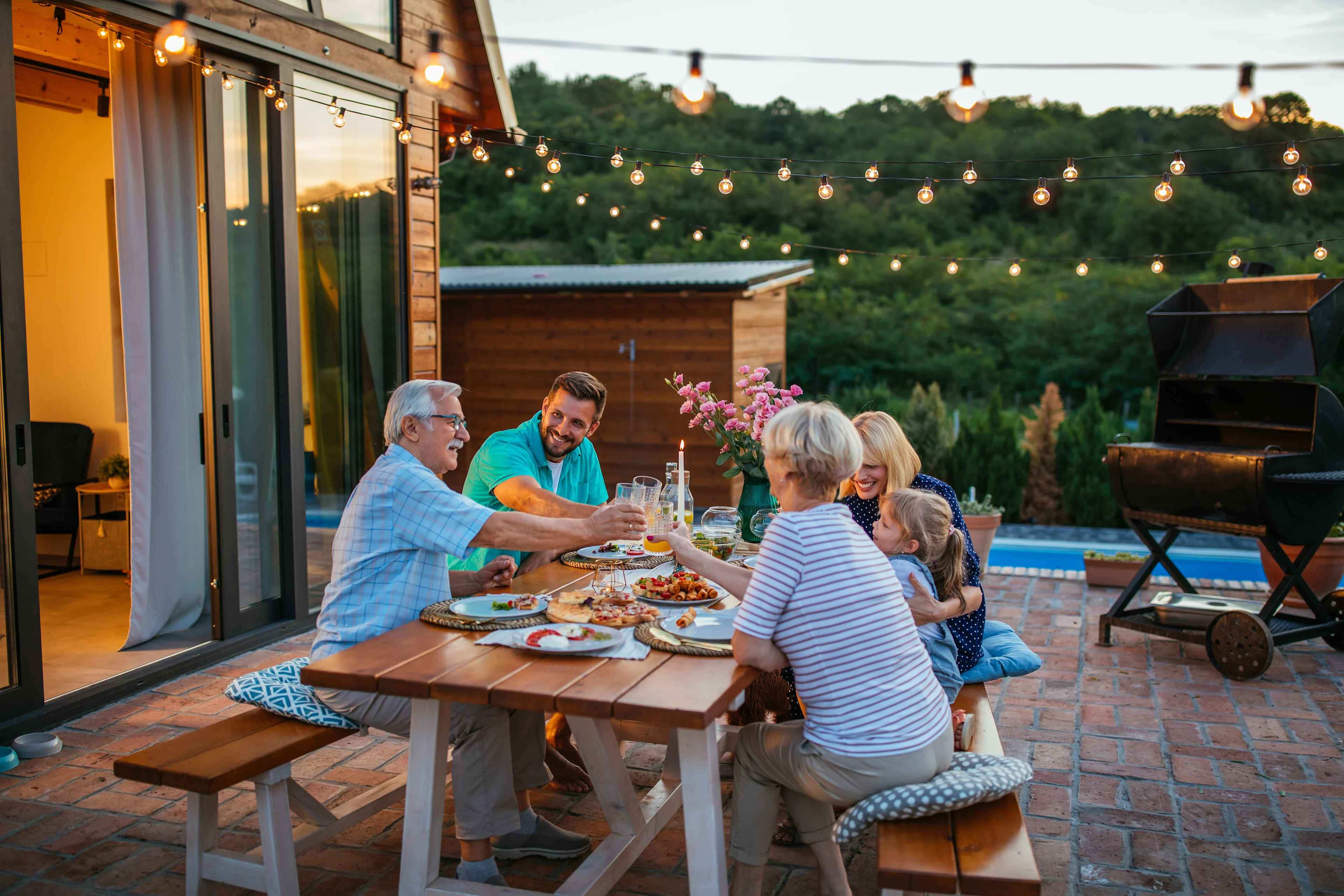 Multi-generational family enjoying outdoor dinner on brick patio with string lights, pool, and grill in background at dusk.