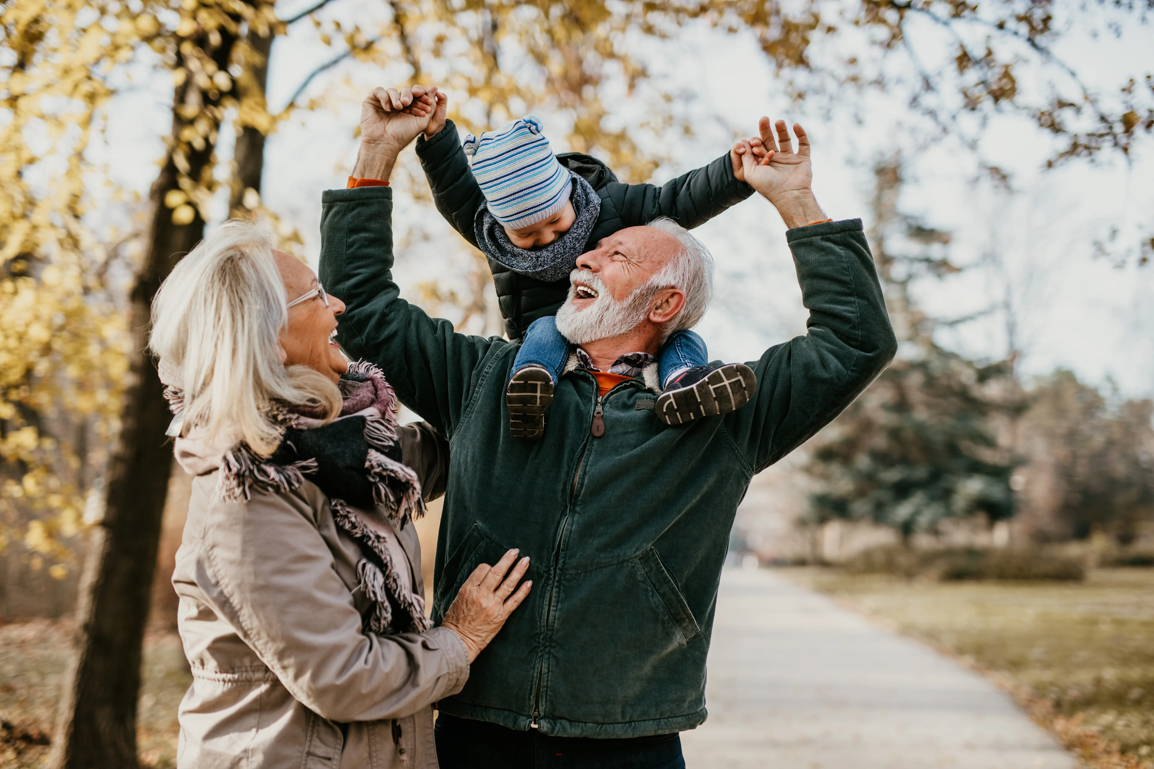 Smiling grandparents playing with grandchild outdoors in autumn park, grandfather carrying child on shoulders.