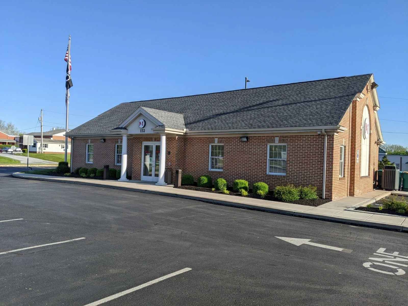 Red brick building with white columns and gray roof, American flag on pole, parking lot in front, clear blue sky.