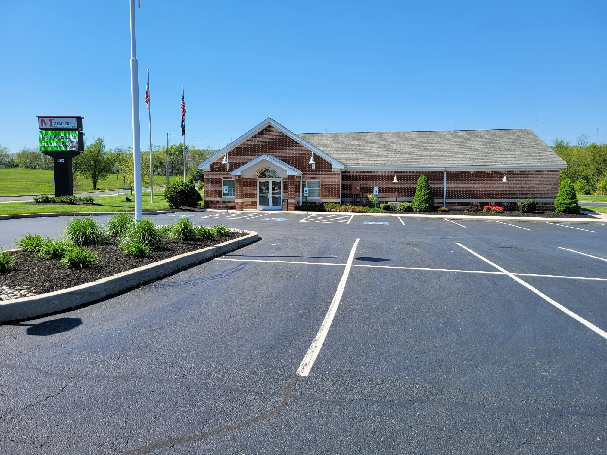 Enola Members 1st Federal Credit Union building with gray roof and white trim, parking lot in foreground, flagpoles and digital sign on left, under clear blue sky.