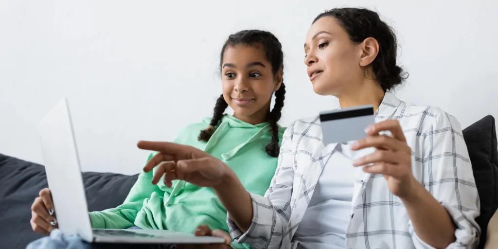 Mother and daughter sitting together with laptop, woman holding credit card while girl points at screen during online shopping.