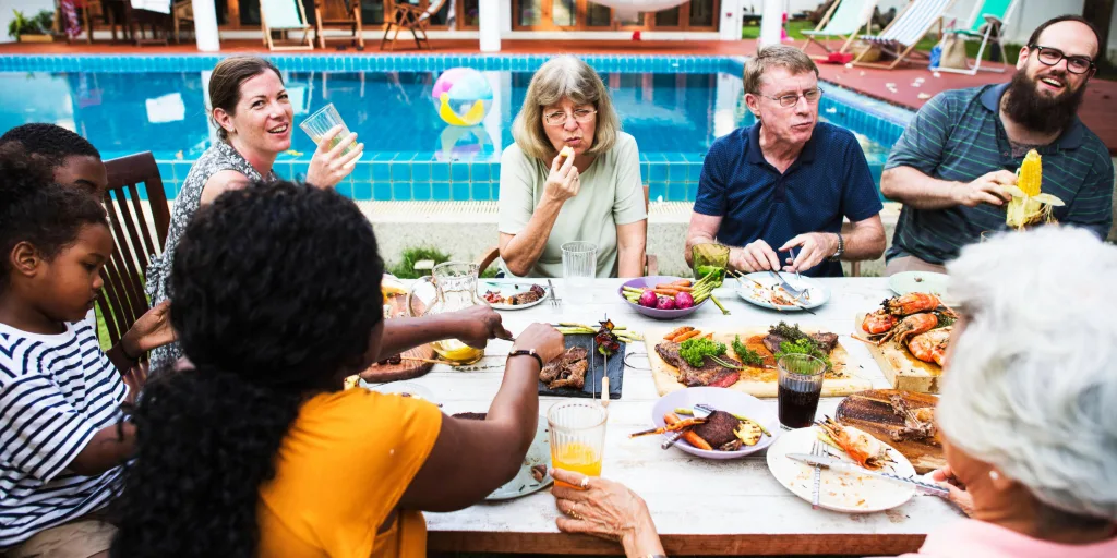 A group of people enjoying a meal together at an outdoor table by a swimming pool with various dishes spread out.