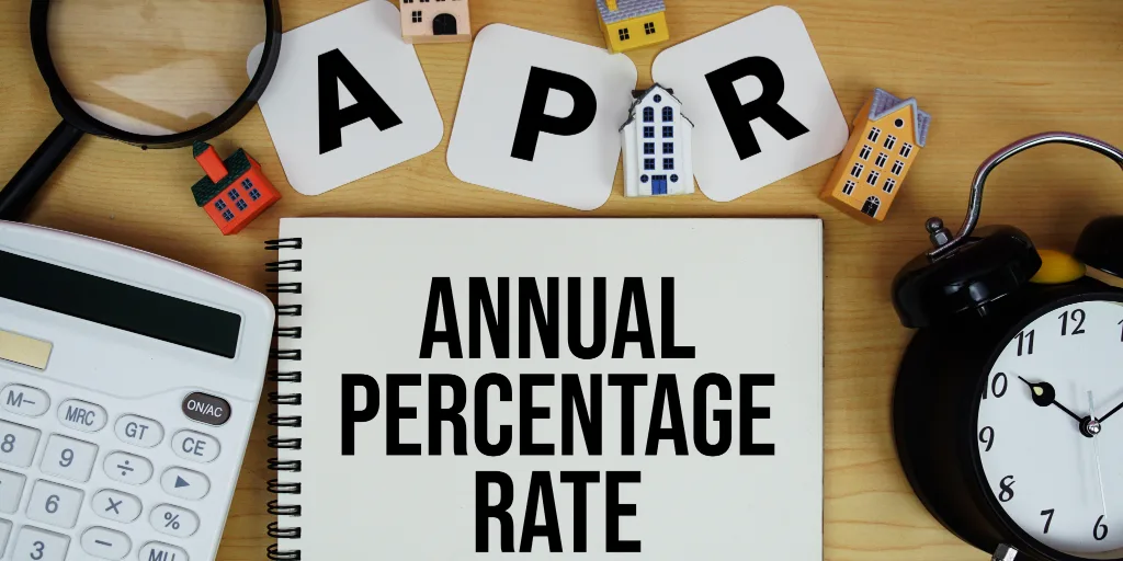 Notebook displaying "ANNUAL PERCENTAGE RATE" with APR letter tiles, miniature houses, calculator, magnifying glass, and clock on wooden desk.