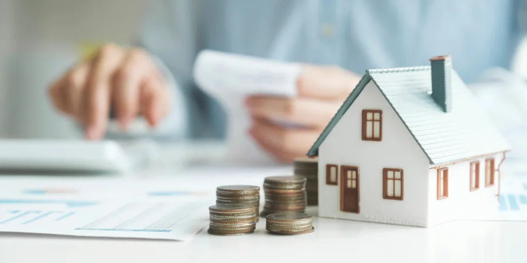 Miniature house model with stacked coins in front, person using calculator in background, representing home financing.