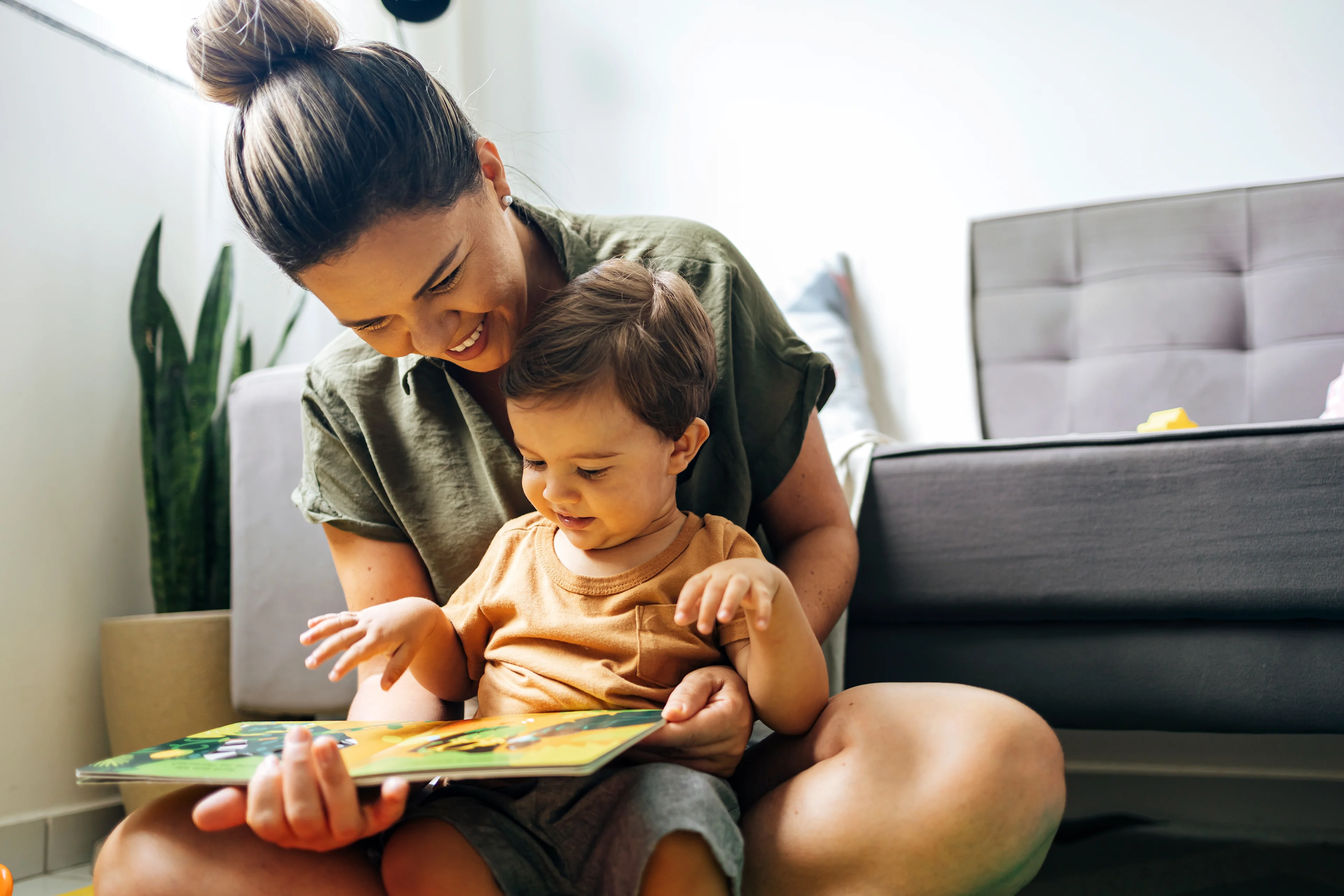 Mother in olive green shirt reading colorful picture book with young child in orange shirt sitting on her lap at home.