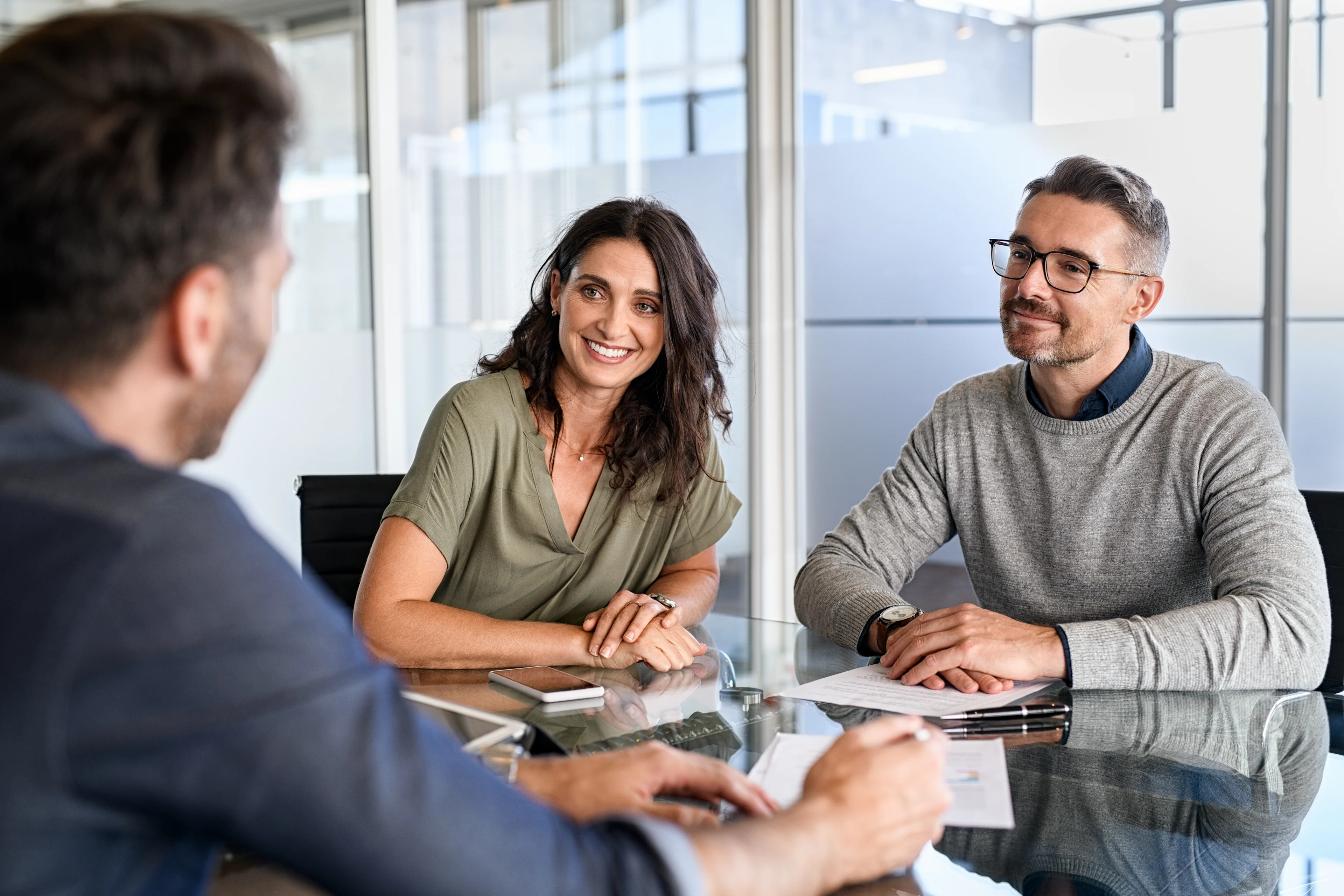 A couple in business meeting at glass table in modern office, woman in olive top and man in sweater smiling at an advisor.