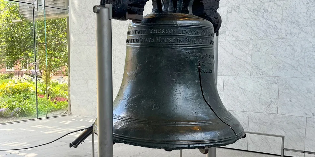 The Liberty Bell on display with its famous crack visible, shown against a white marble wall with greenery visible outside.