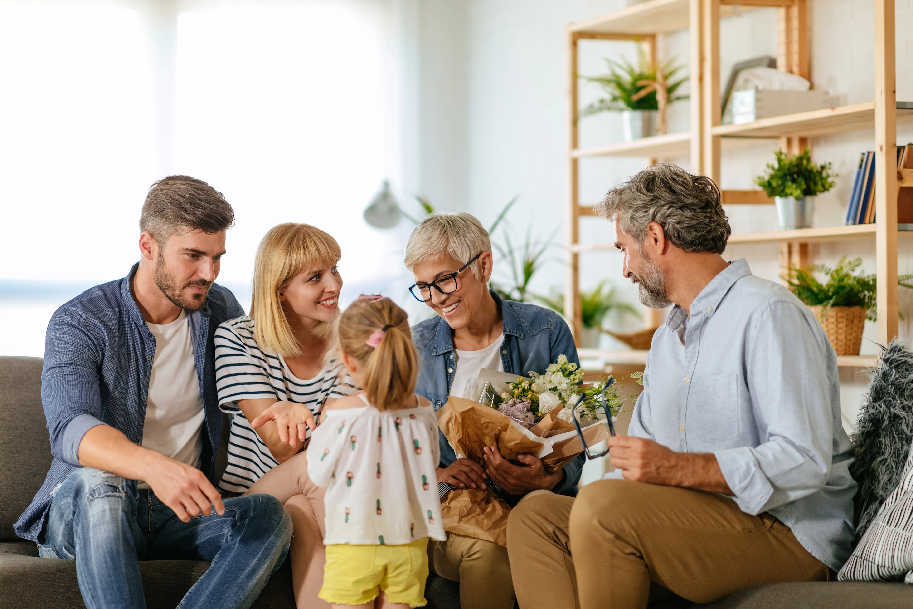 Family gathering in bright living room with young girl presenting flowers to smiling grandmother while parents and grandfather look on.