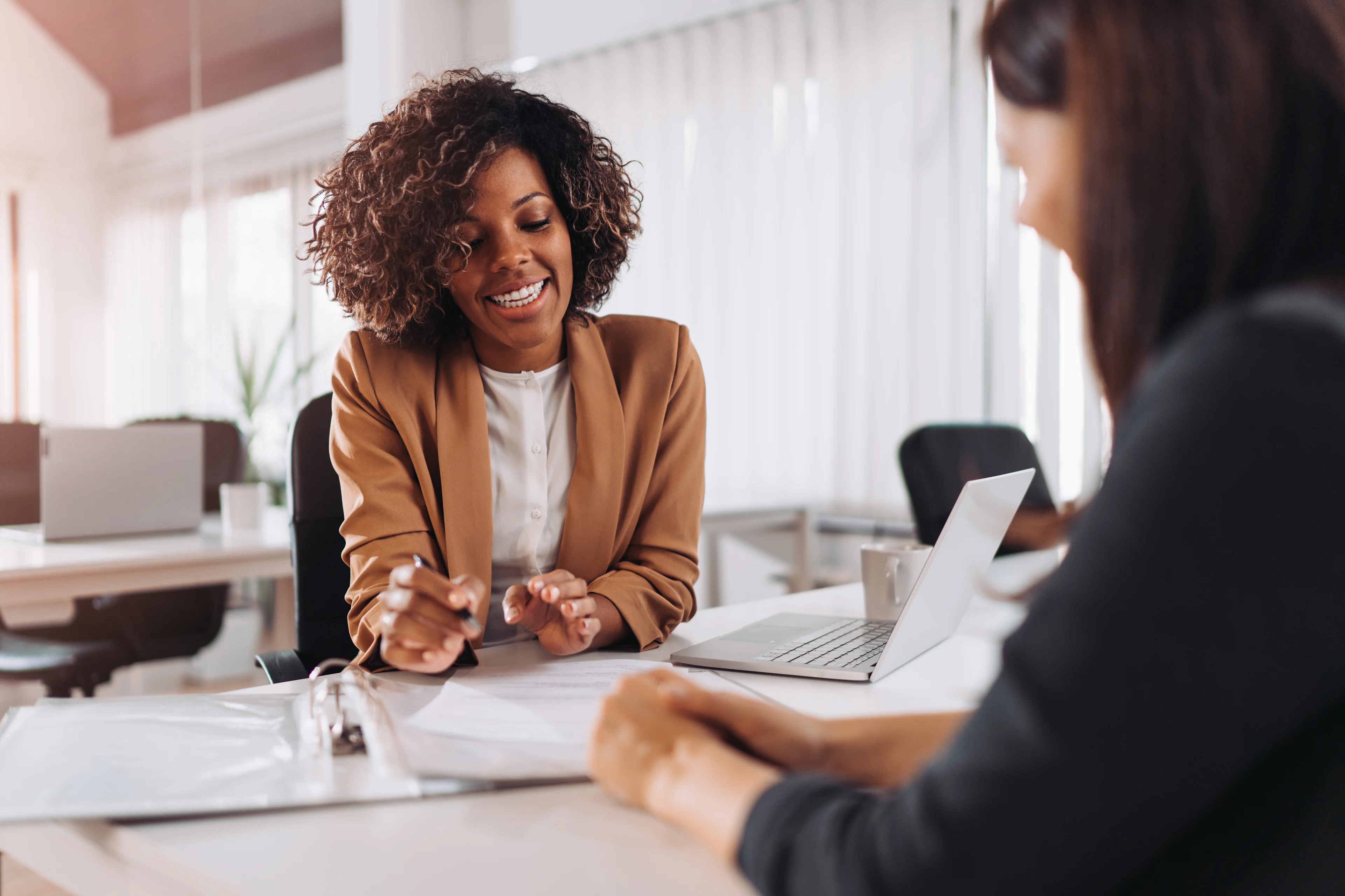 Woman in tan blazer smiling while discussing documents with colleague at desk with laptop in bright office setting