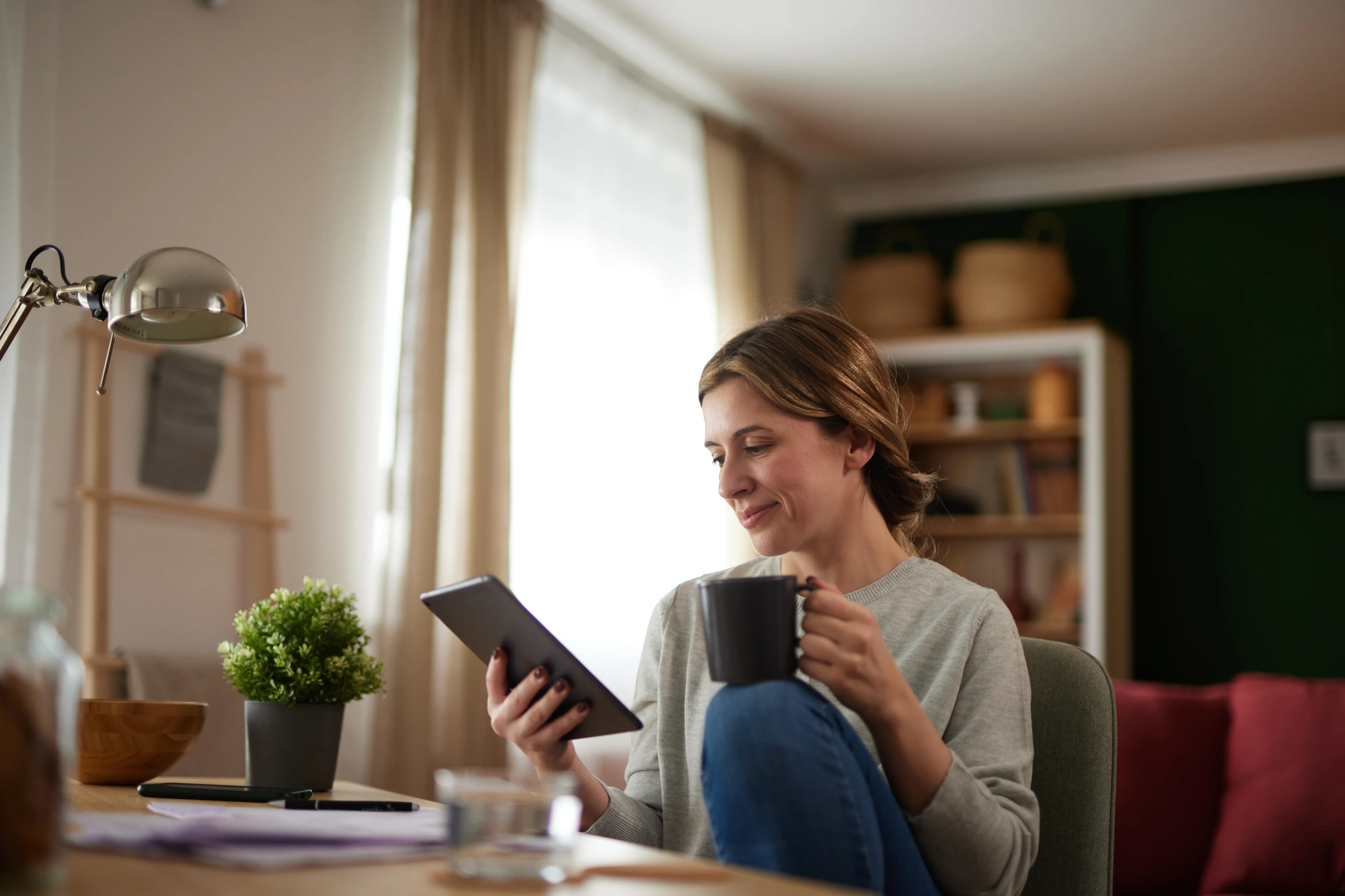 Woman in gray sweater relaxing at home, holding tablet and coffee mug while sitting in chair near bright window and desk lamp.