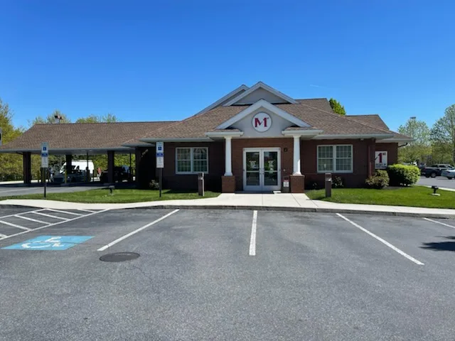 Single-story brown brick commercial building with covered entrance, peaked roof with M logo, and parking lot under blue sky
