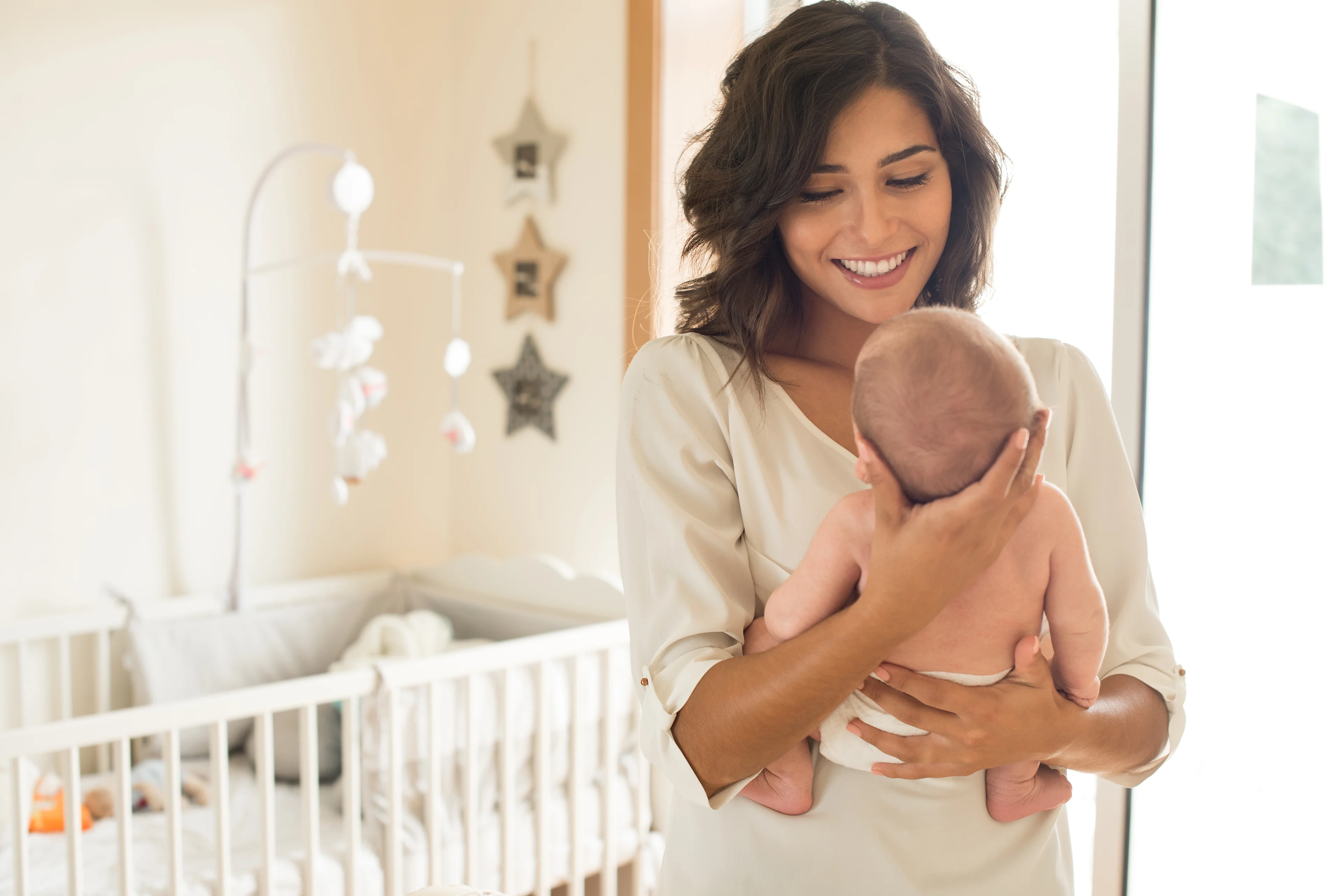 Smiling mother in cream blouse holding newborn baby in bright nursery with white crib and star decorations.