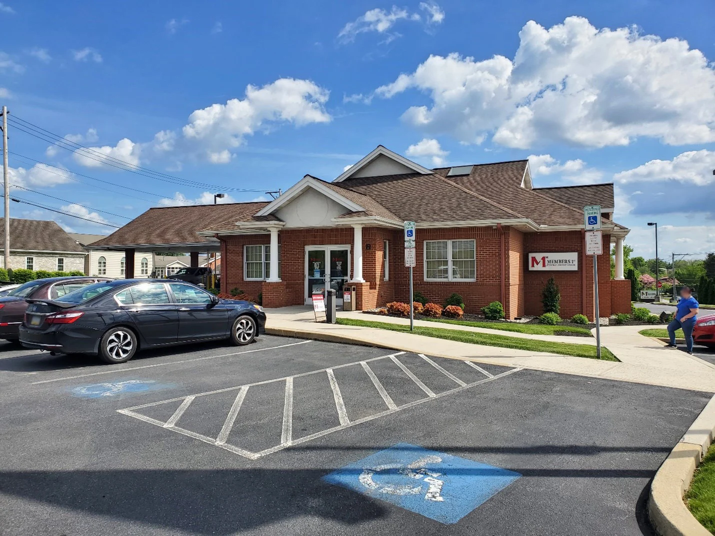 Red brick bank building with white columns and brown roof under blue sky with white clouds and parking lot in foreground.