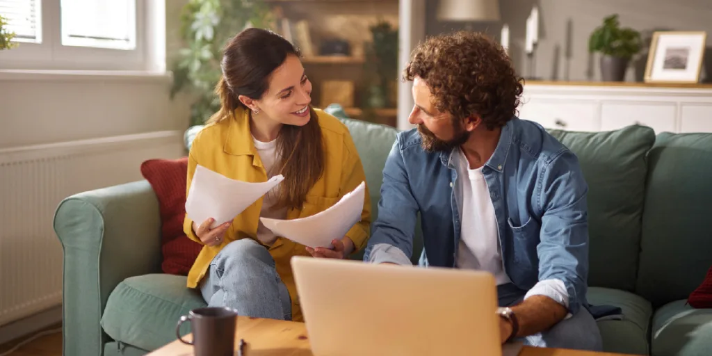 Couple sitting on green couch reviewing documents together with laptop, woman in yellow cardigan, man in blue denim shirt.