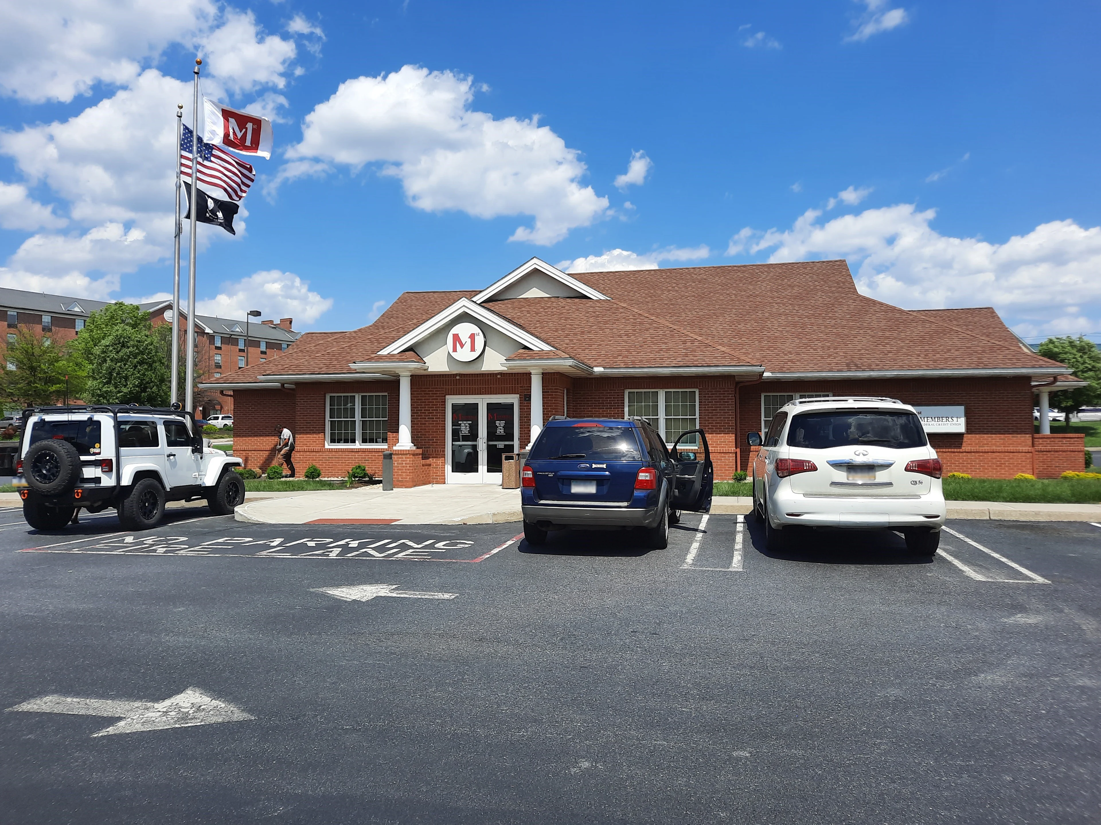 Red brick building with white columns and M logo, three flags flying, parking lot with vehicles under blue sky with clouds.