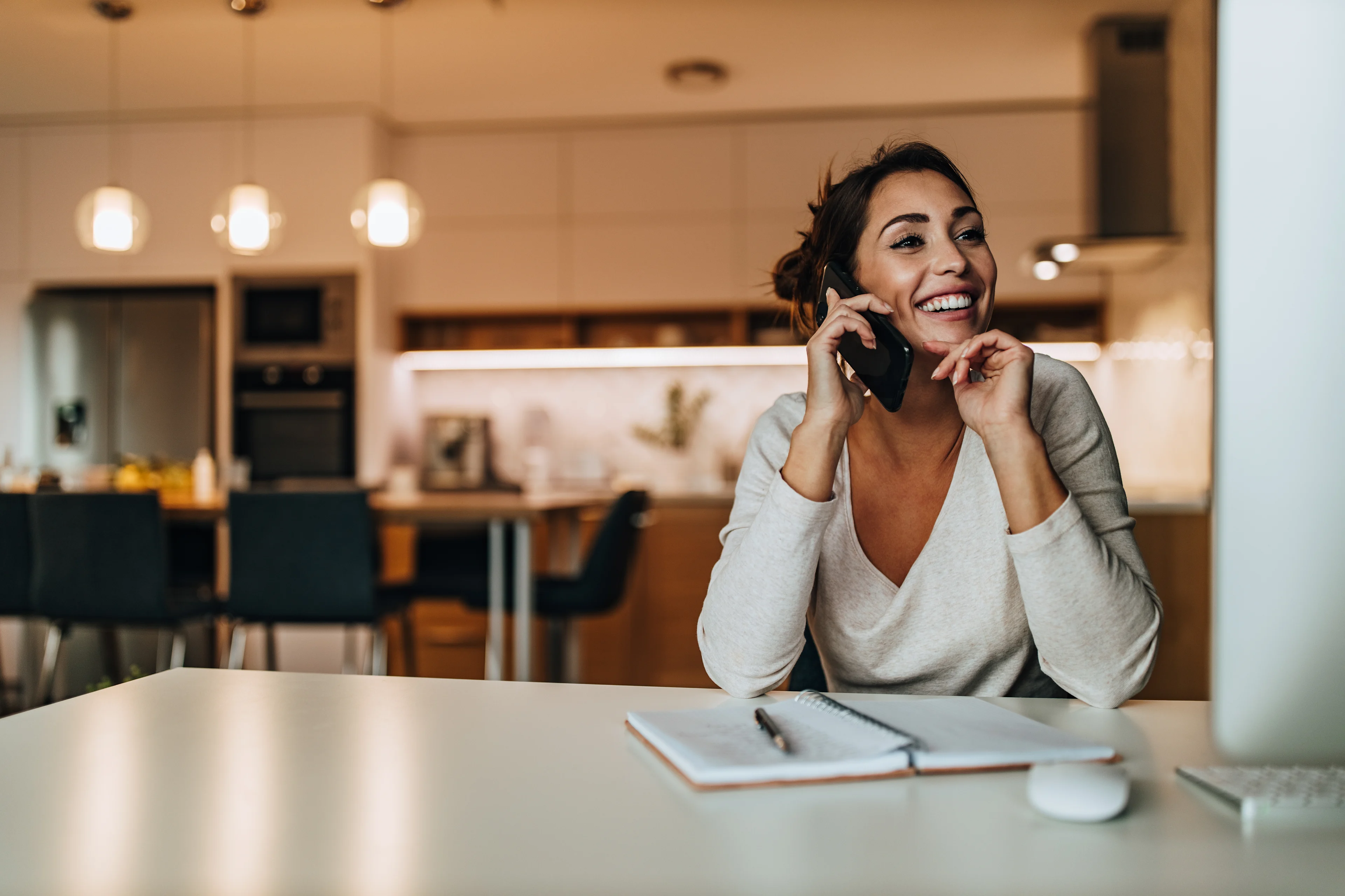 Smiling woman in gray sweater talking on phone at home office desk with notebook and laptop in modern kitchen setting.