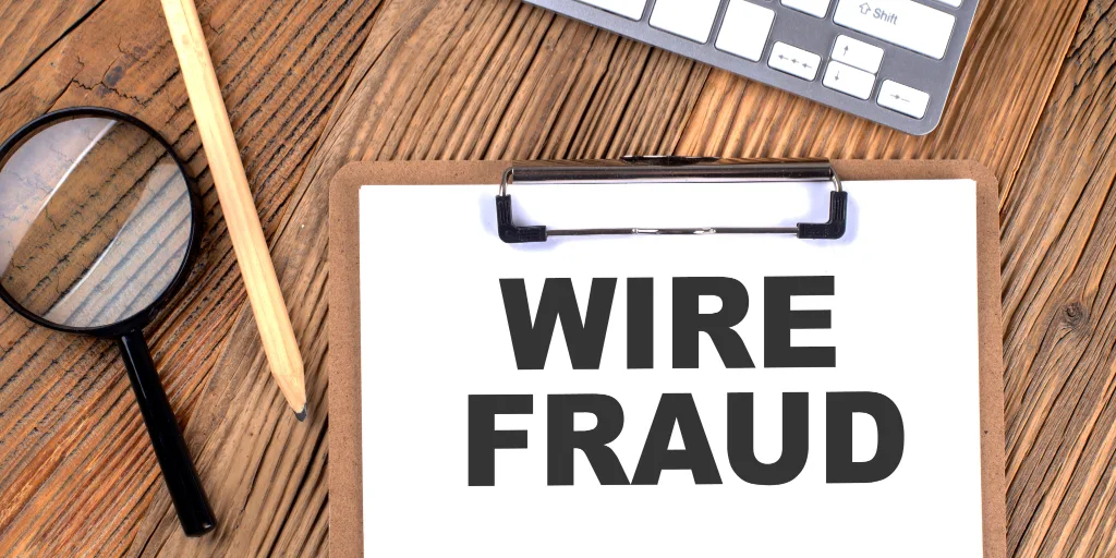 Clipboard with WIRE FRAUD text on wooden desk with magnifying glass, pencil, and keyboard visible