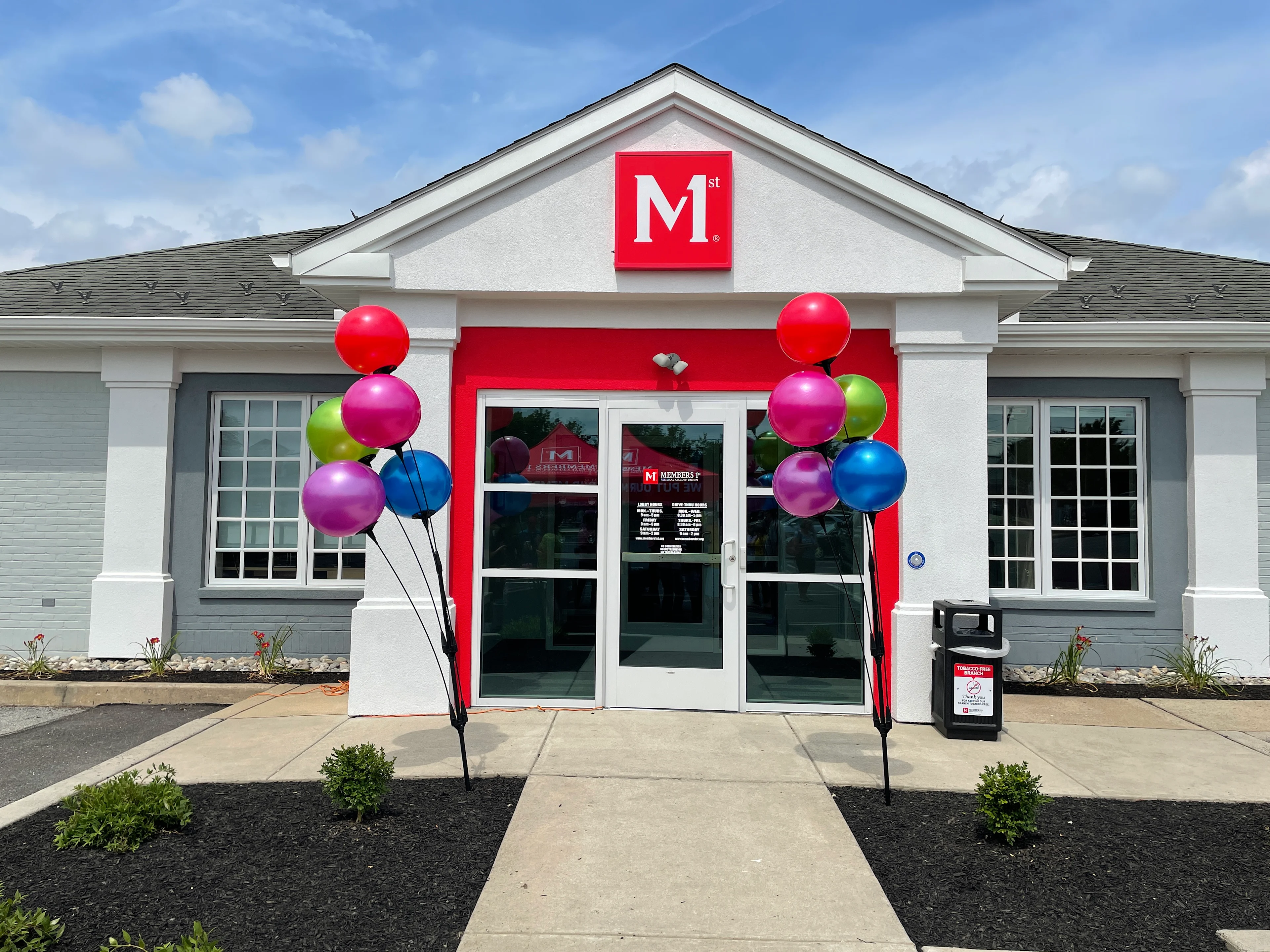 Macungie Members 1st Federal Credit Union branch white and gray building with red M logo and bright red entrance frame, decorated with colorful balloons on both sides.