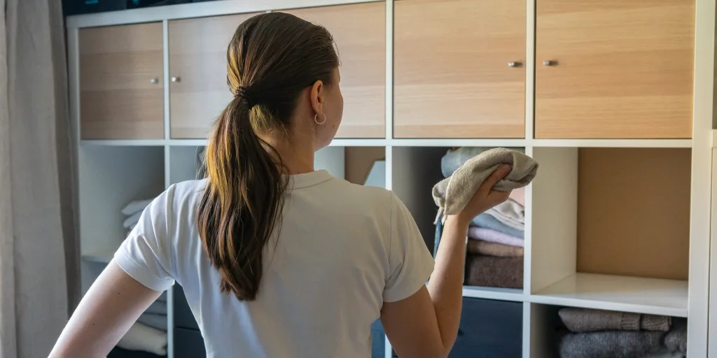 Woman with ponytail in white shirt organizing folded towels in wooden storage cubby shelving unit.