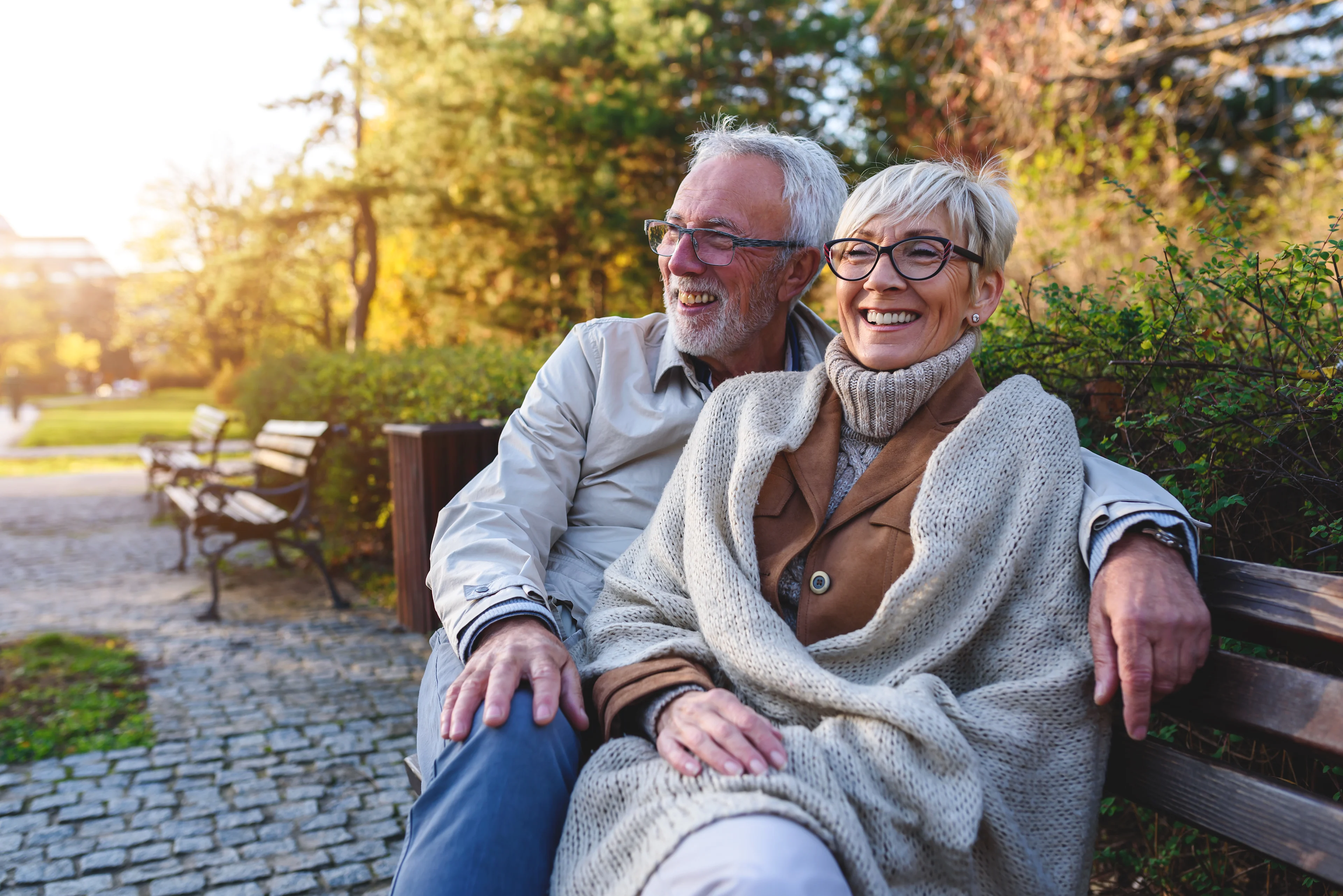 Happy senior couple wearing glasses sitting together on park bench in autumn, smiling and embracing outdoors.