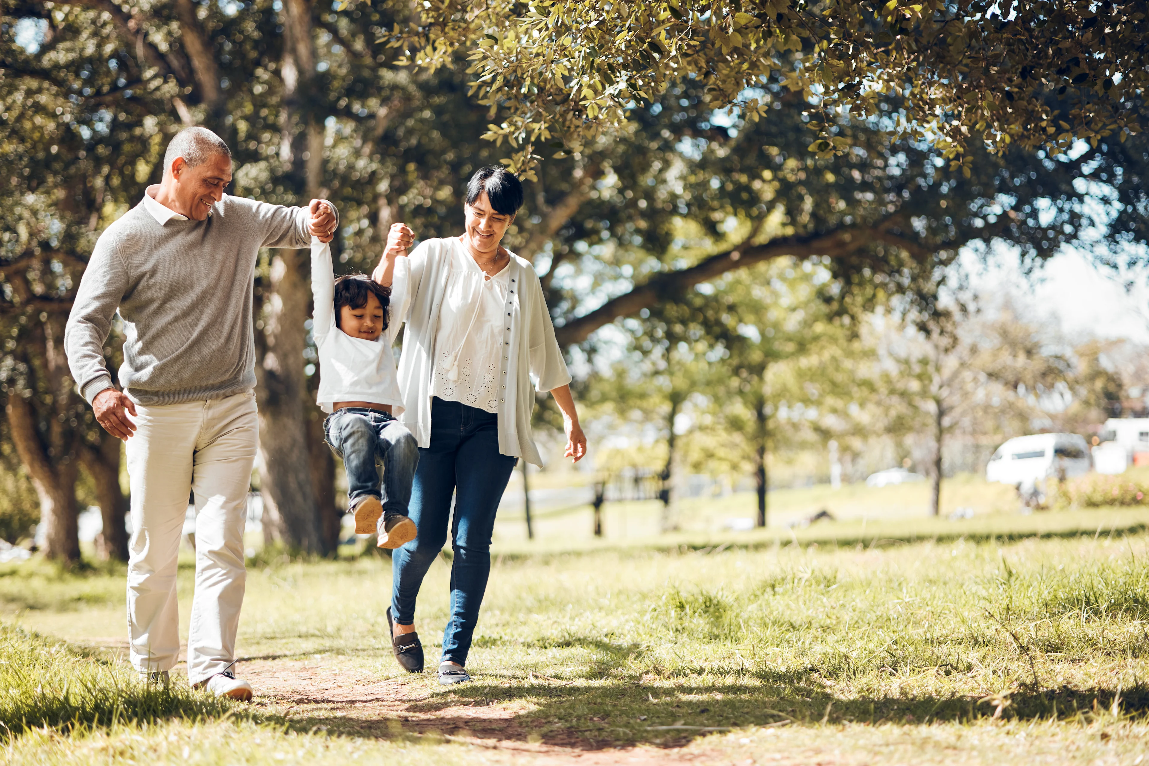 Grandparents swinging young child between them while walking on sunny park path surrounded by green trees and grass.