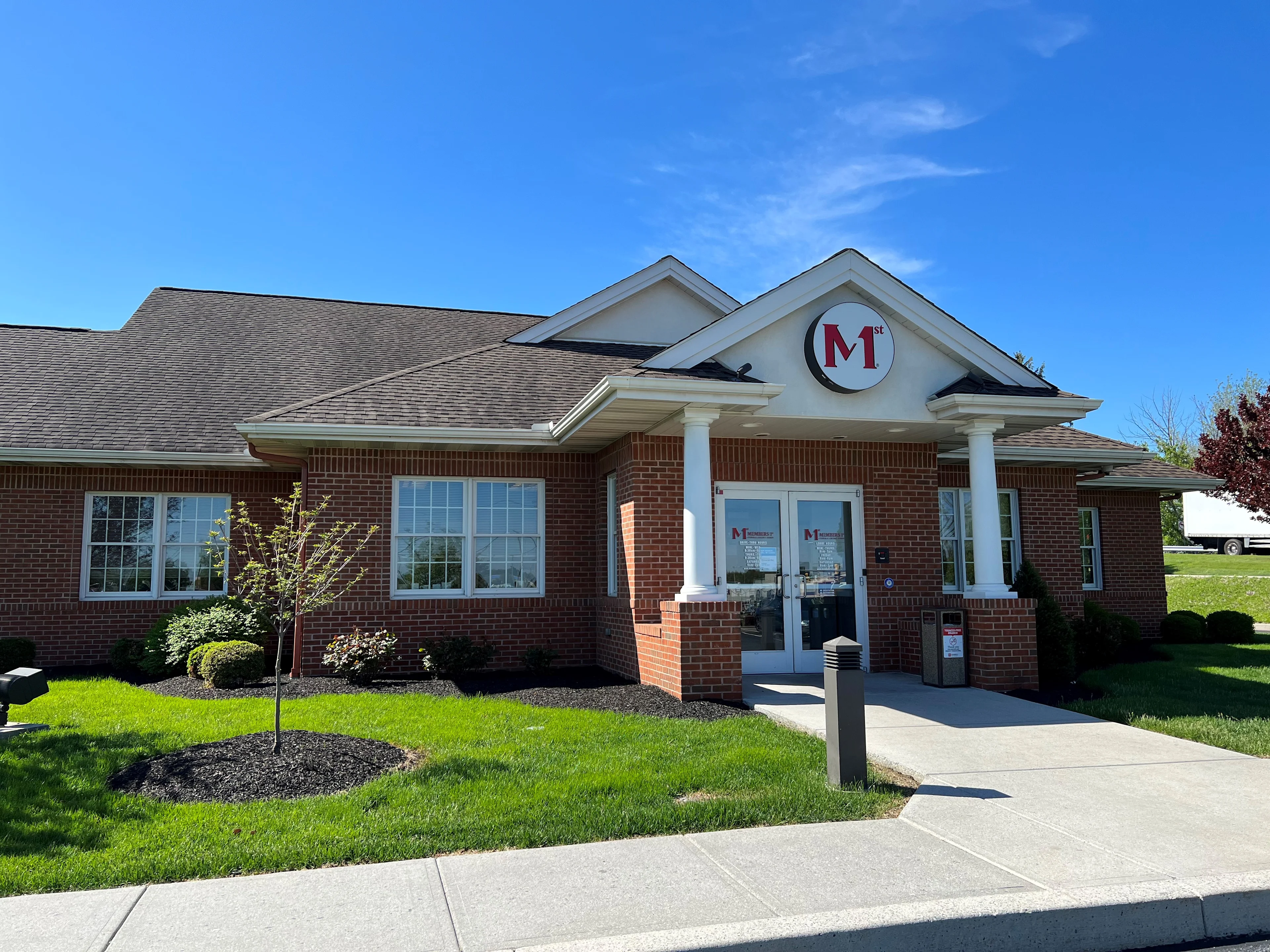 Red brick building with white columns and M1st logo above entrance, green lawn and landscaping, clear blue sky.