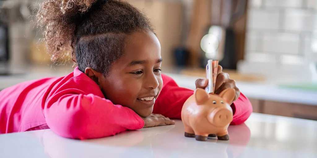 Young girl in bright pink shirt smiling while inserting coin into piggy bank on white counter, learning about saving money.