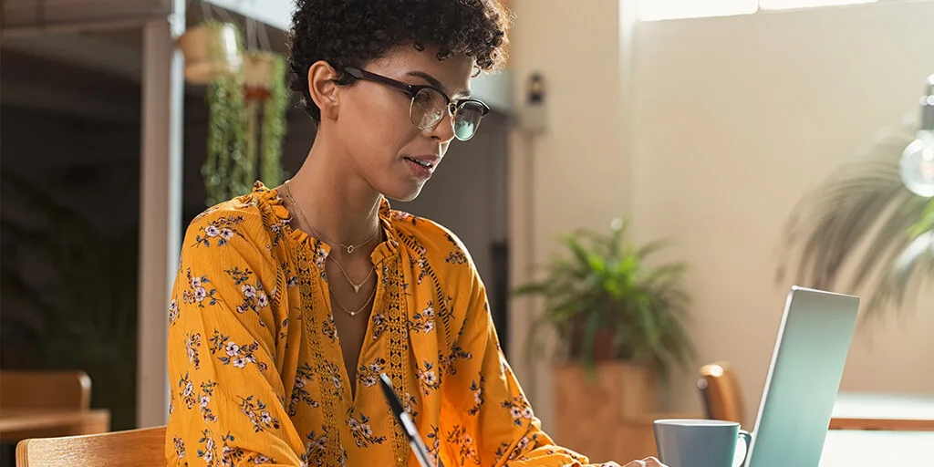 Woman with curly hair and glasses wearing yellow floral blouse working on laptop at wooden table with coffee cup nearby.