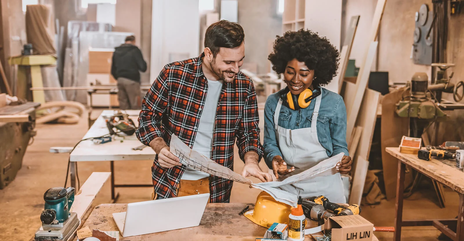 Two smiling people reviewing blueprints in a woodworking workshop with tools and materials on workbench.