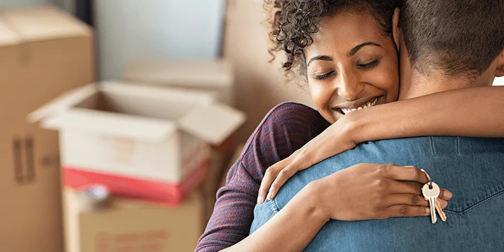 Smiling woman hugging man while holding keys, with moving boxes in background, suggesting a new home celebration.