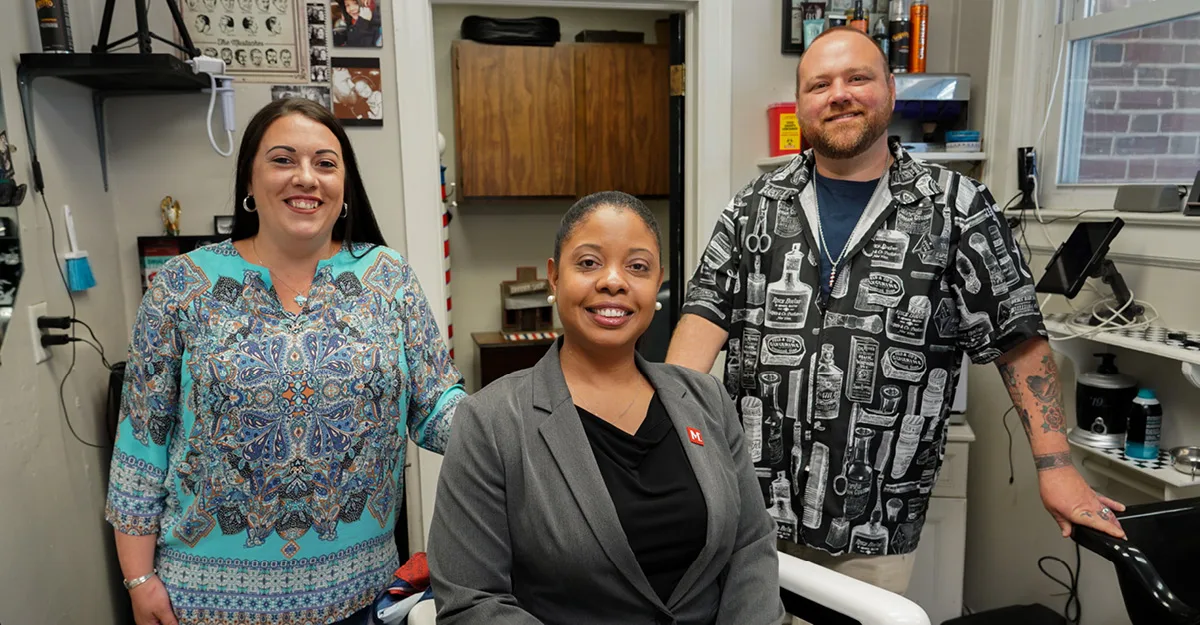 Three smiling healthcare workers in colorful patterned scrubs standing together in a medical office with equipment visible.