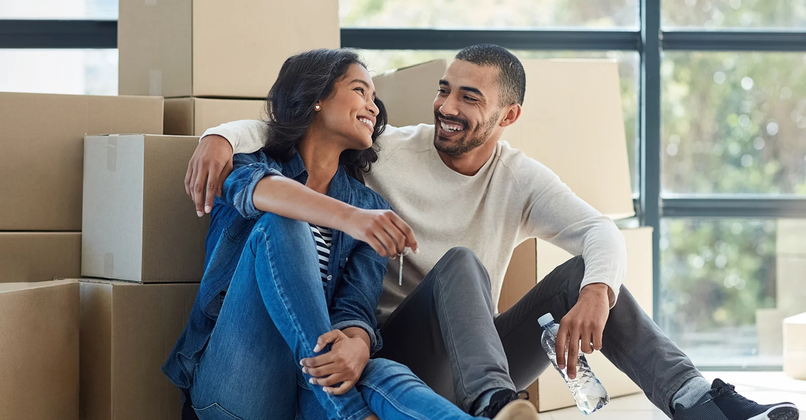 Happy couple sitting on floor with moving boxes, holding keys to their new home, smiling at each other.