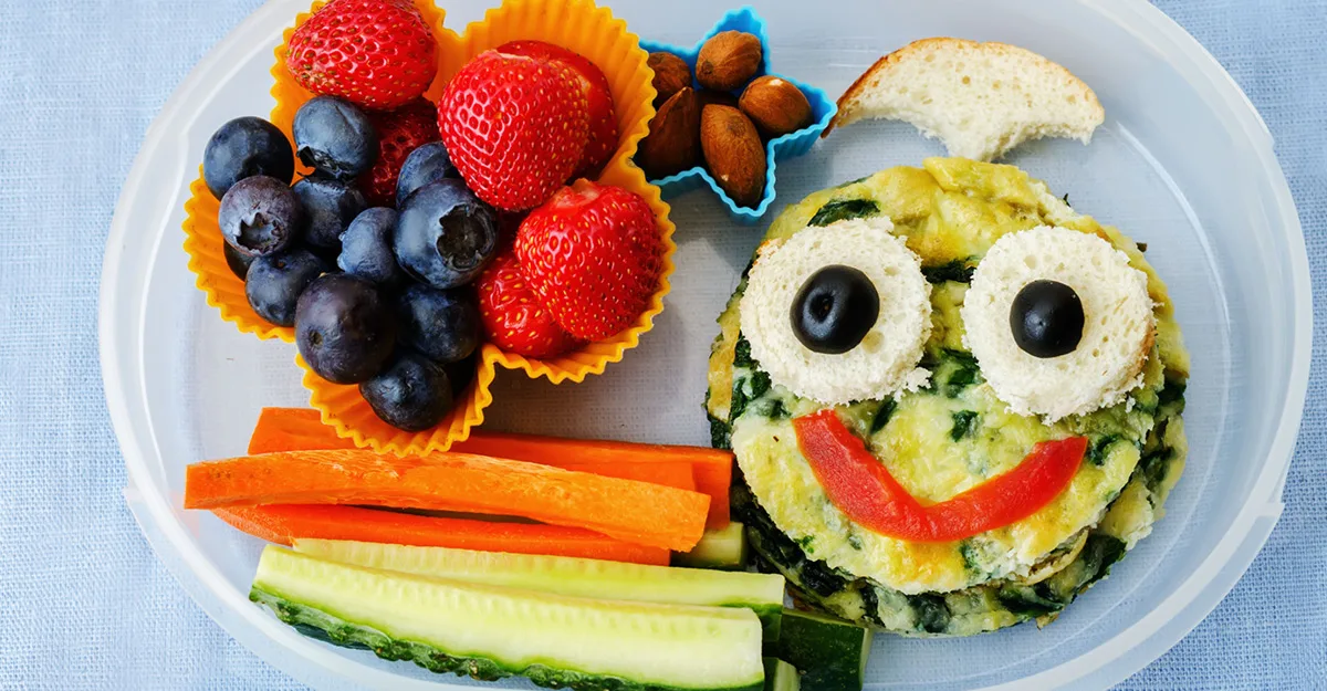 Creative kids lunch plate with smiling face made from eggs, bread, and vegetables, plus berries, nuts, and fruit.
