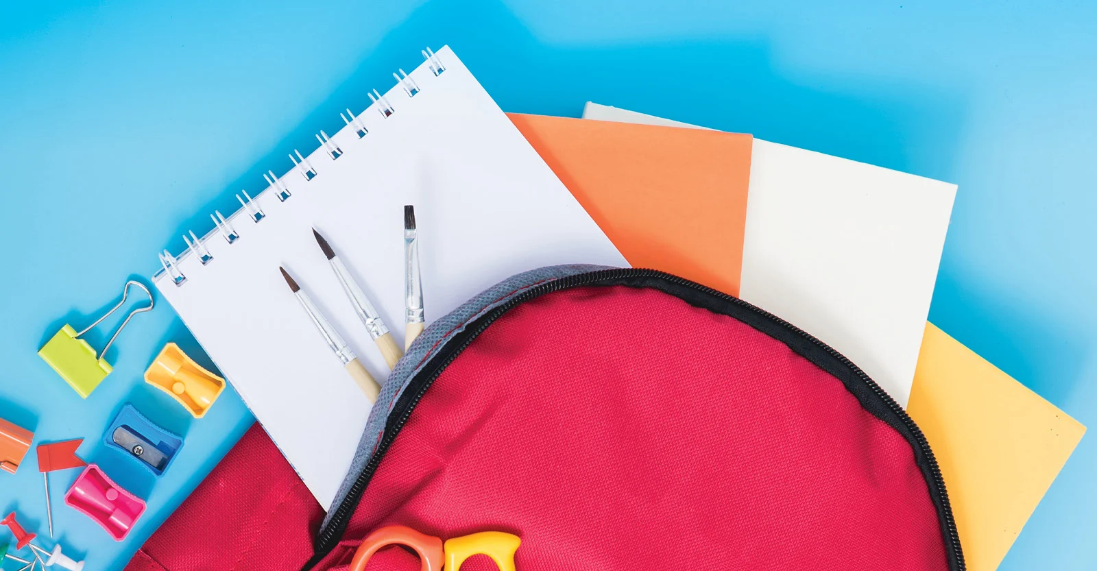 Red backpack with school supplies including notebooks, pens, and colorful sharpeners on a bright blue background.