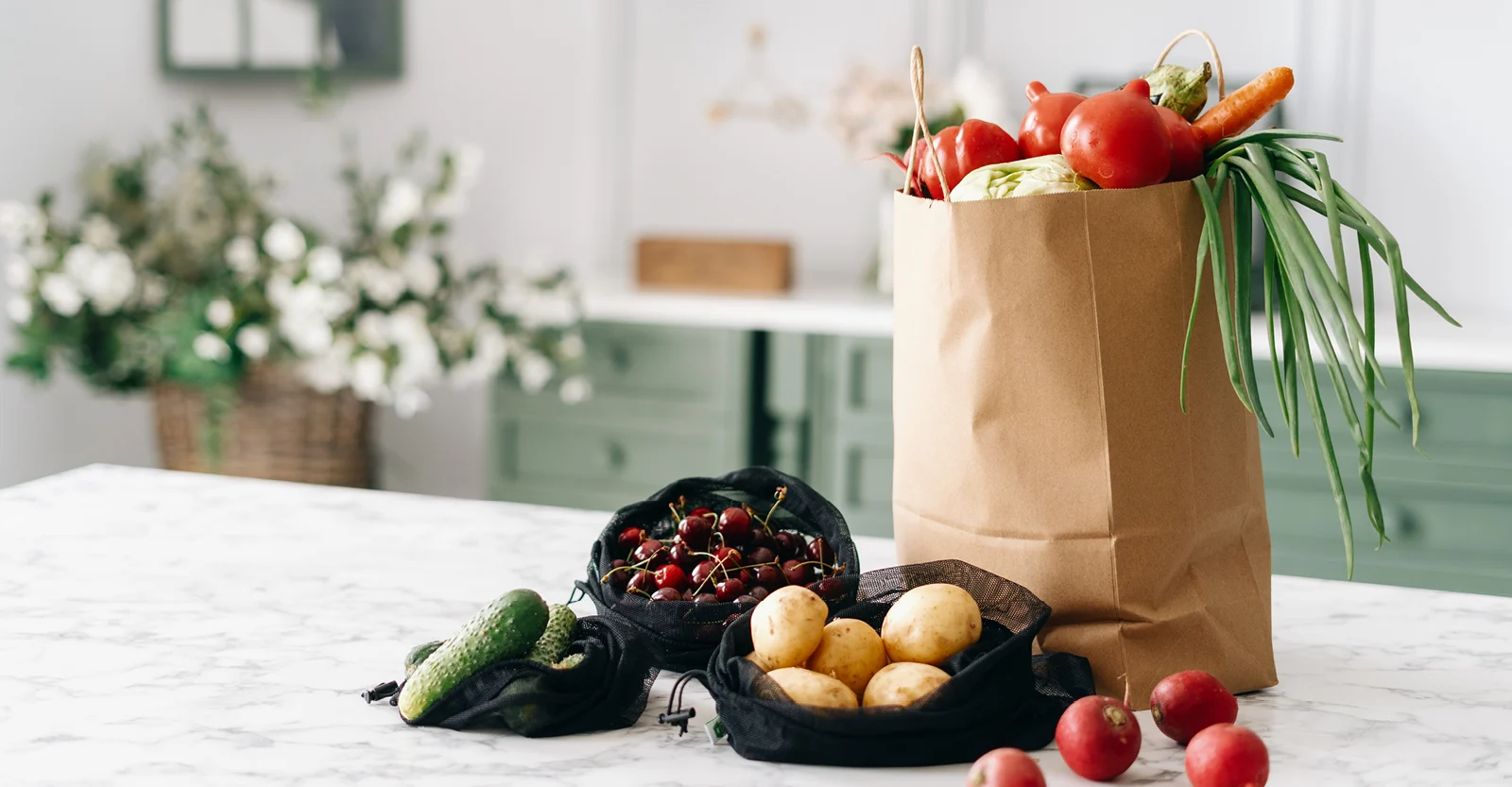 Brown paper grocery bag filled with fresh vegetables including red peppers, carrots, and leeks on a white kitchen counter.