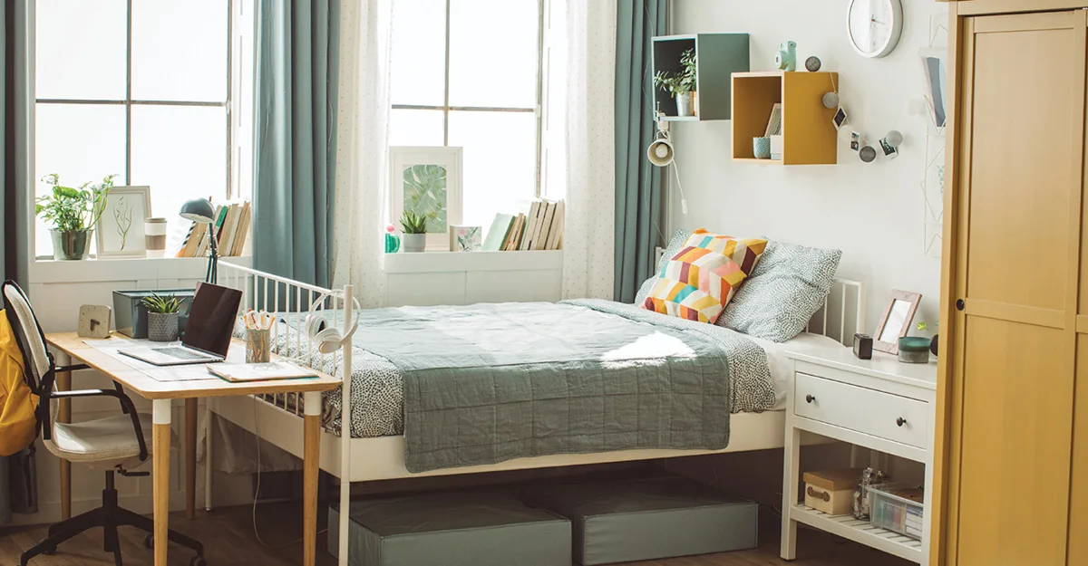 Bright bedroom with white bed, sage green bedding, colorful pillows, desk with laptop, plants, and blue curtains.
