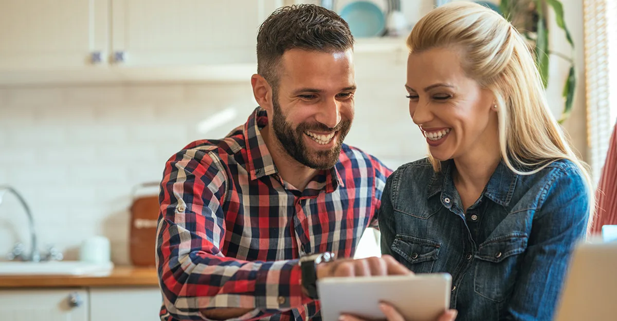 Happy couple smiling while looking at a tablet together in a bright modern kitchen with white cabinets.