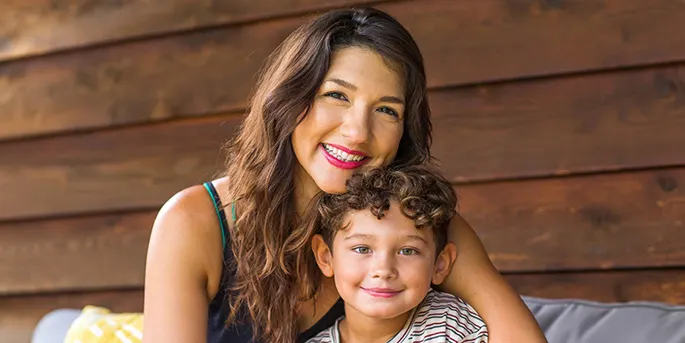 Smiling woman with long brown hair and red lipstick embracing a young curly-haired child against a wooden wall background.