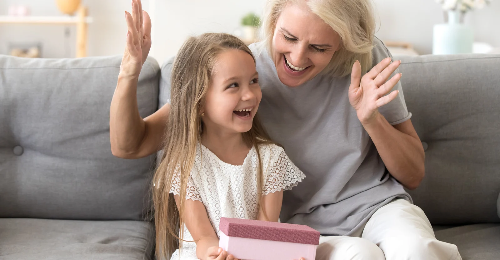 Smiling grandmother and granddaughter giving high-five while sitting on gray couch with pink gift box in bright home.