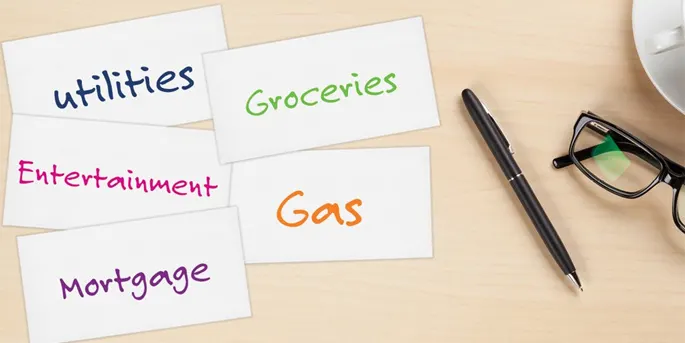 Index cards labeled with budget categories including Utilities, Groceries, Entertainment, Gas, and Mortgage on a desk.