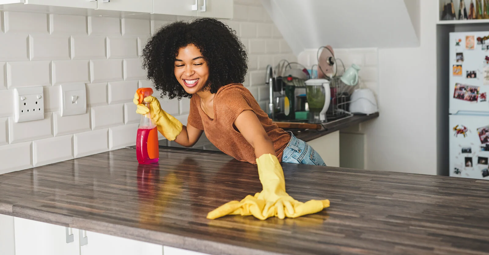 Smiling woman wearing yellow gloves cleaning kitchen counter with spray bottle and cloth in modern kitchen