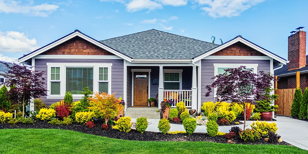 Gray single-story house with white trim, covered porch, and colorful landscaping with yellow and red shrubs under blue sky.